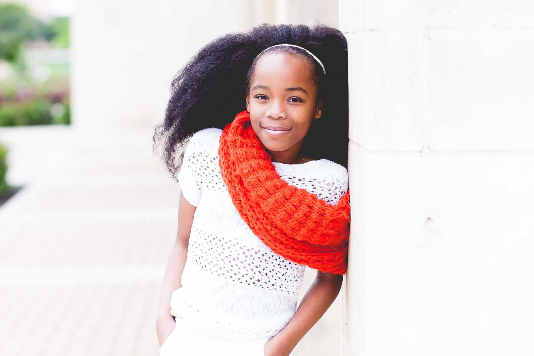 A young girl with voluminous natural hair, wearing a white crochet top and a bright red scarf, leans against a white pillar while smiling confidently.