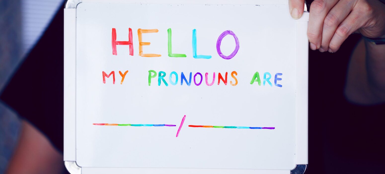 A person holds a whiteboard with colorful text reading "HELLO MY PRONOUNS ARE" followed by a blank line. The board's edge is visible, and the person's hand is partially shown.