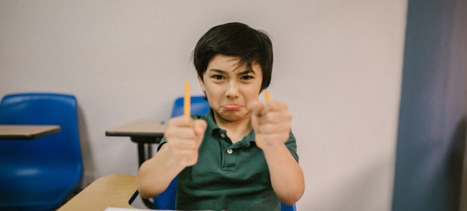 A young boy sitting at a desk in a classroom, holding two pencils with a playful, pouty expression, while blue chairs and a notebook are visible in the background.