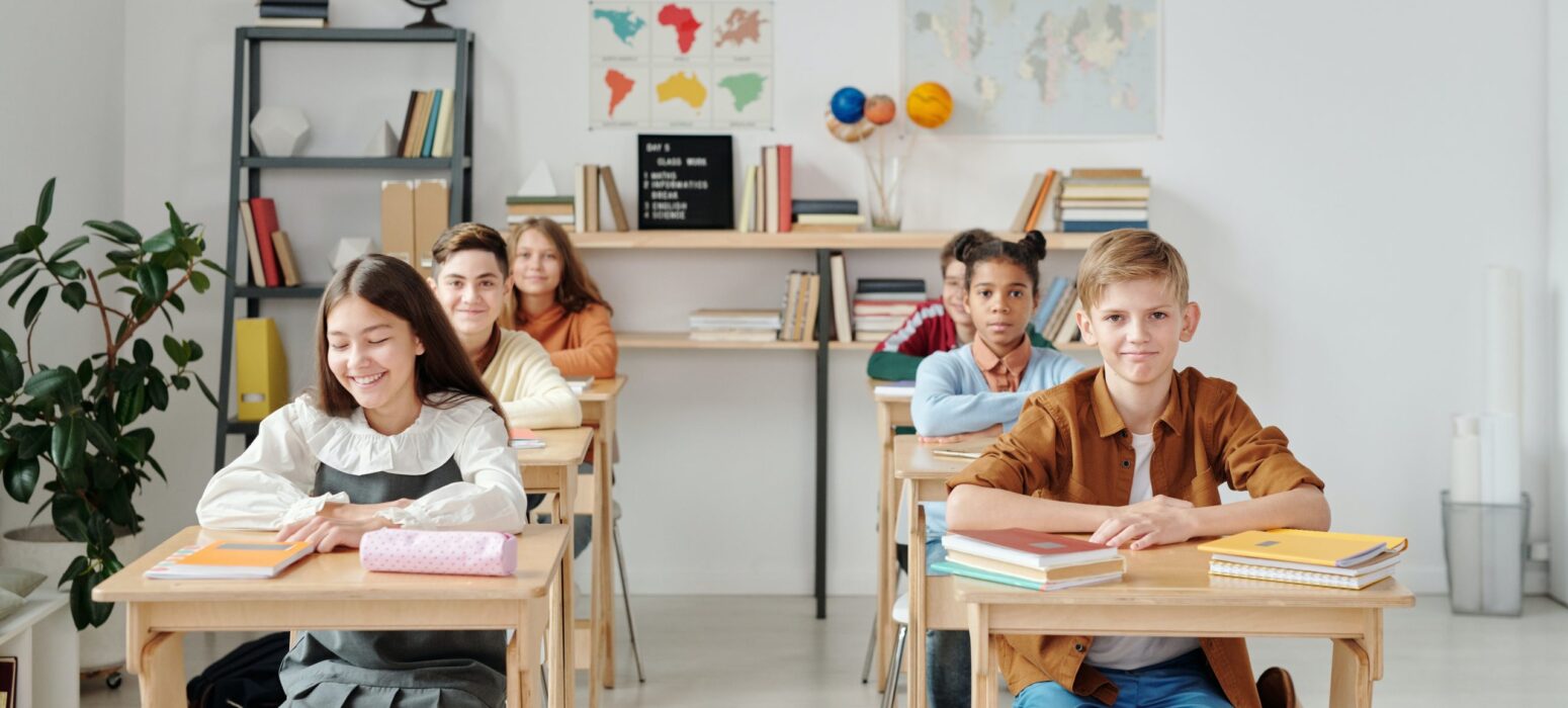 A classroom of diverse students sits attentively at their desks, smiling and engaged. The room is bright and well-organized, with shelves of books, educational globes, and maps adorning the walls. The students appear to be in a positive and focused learning environment.