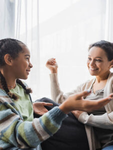 teenage african american girl pointing with hand while talking to smiling mother on sofa