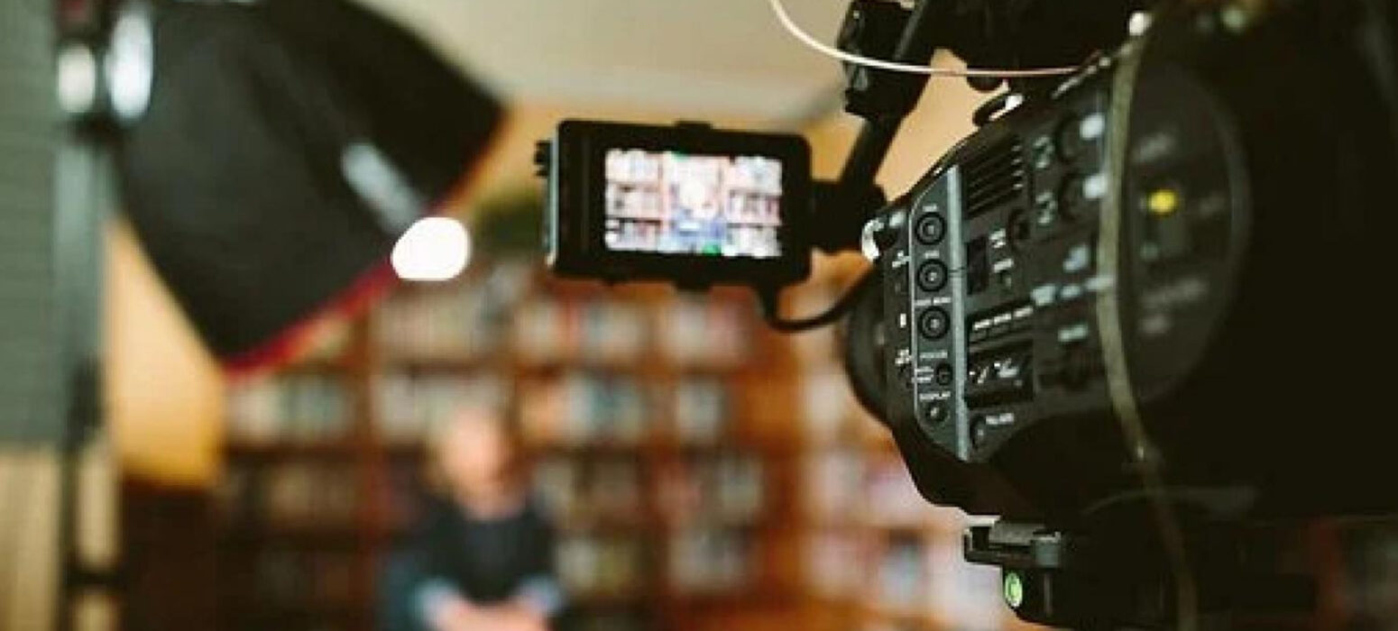 A professional video camera records an interview in a library setting, capturing a blurred person seated in the background while the camera’s screens display a clearer view of the subject.