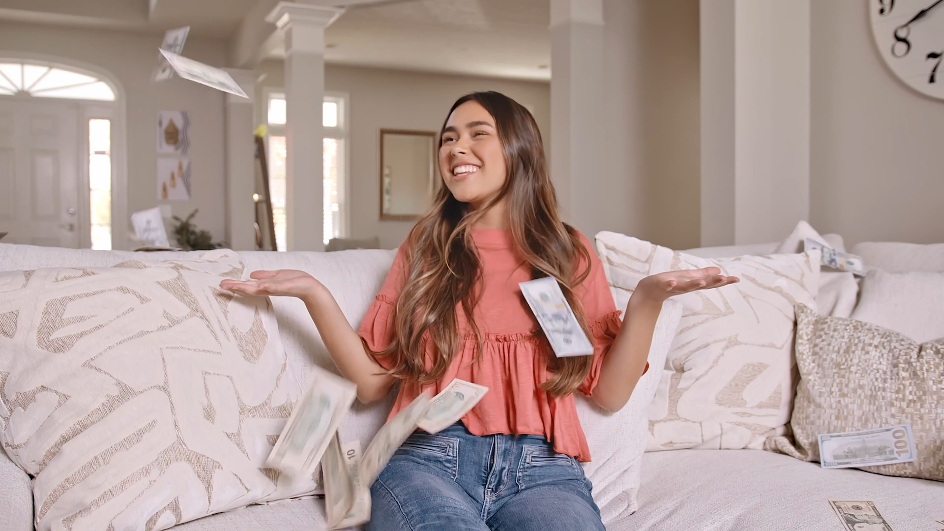 The image depicts Babysitter Boss, Sophia. She is sitting on a light-colored sofa in a cozy and well-lit living room. She is wearing a coral-colored top with ruffled sleeves and jeans. She has an excited expression, smiling broadly, with her arms open as dollar bills appear to fall around her, emphasizing a sense of financial reward or abundance. The background features decorative pillows, a clock on the wall, and soft neutral tones, contributing to a relaxed and positive atmosphere. The scene suggests excitement about earning or financial success from babysitting jobs.