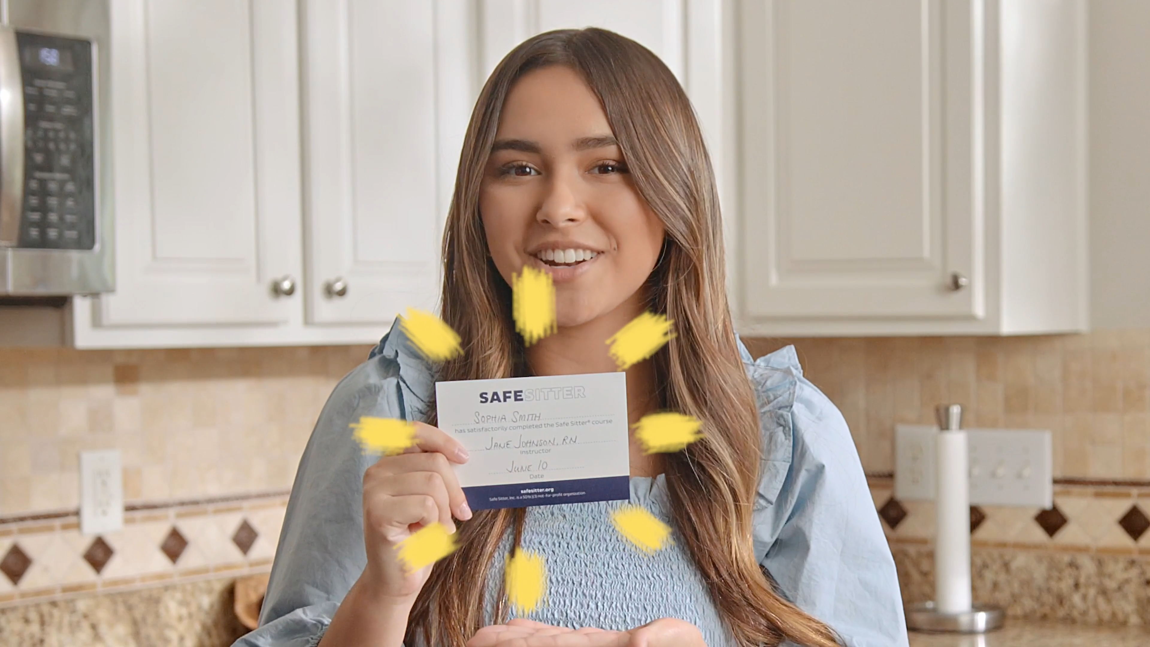 The image shows Babysitter Boss, Sophia, smiling and holding a certificate that reads "Safe Sitter." She is standing in a kitchen with light-colored cabinets and a beige tile backsplash. The certificate, for completing a Safe Sitter course, has handwritten details such as a name, date, and instructor signature. Yellow highlights or burst marks surround the certificate, drawing attention to it. Sophia is dressed in a light blue blouse with ruffled details, and the atmosphere conveys a sense of accomplishment and celebration.