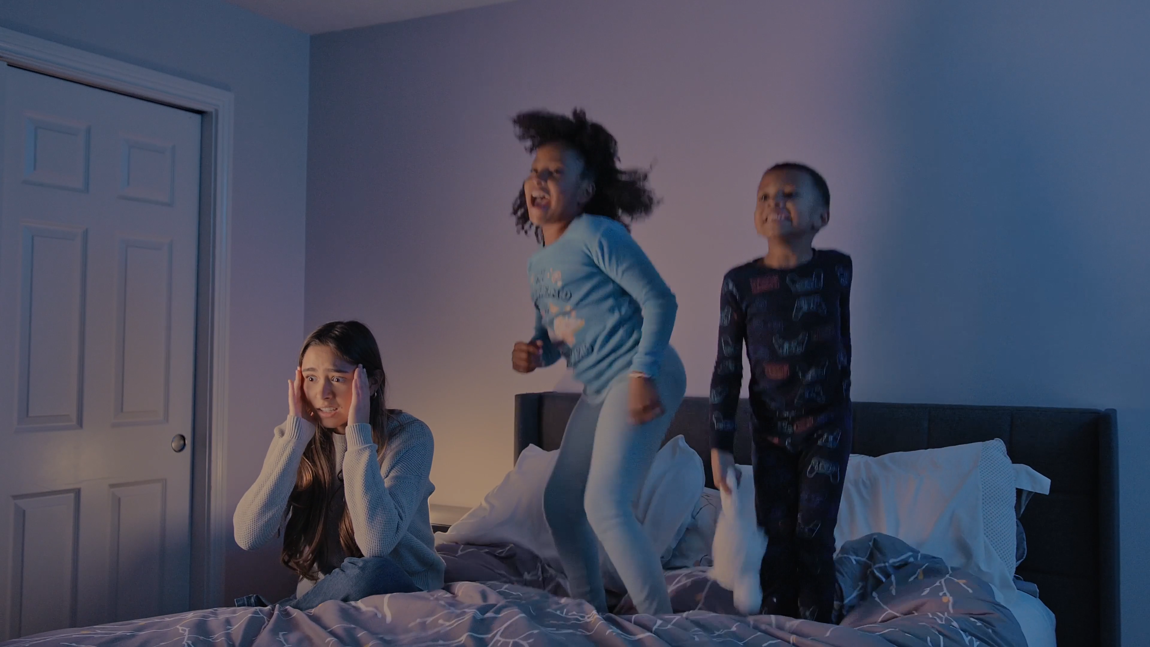 The image shows a chaotic bedtime scene in a dimly lit bedroom. Two children, a girl in blue pajamas and a boy in dark-patterned pajamas, are energetically jumping on a bed with visible excitement and laughter. Babysitter Boss, Sophia, is sitting on the bed and appears overwhelmed, holding her head in her hands, suggesting a sense of frustration or fatigue. The bed has gray and white patterned bedding, and the room's soft lighting creates a cozy, albeit hectic, atmosphere. The scene humorously portrays the challenges of managing active children during bedtime.