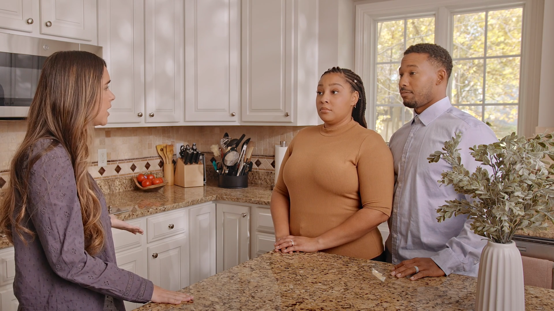 The image shows Babysitter Boss, Sophia, engaging in a conversation with a couple in a bright kitchen. Sophia is on the left, dressed in a purple blouse, and appears to be explaining or discussing something while resting her hands on a granite countertop. The couple, standing on the opposite side of the counter, are listening attentively. The woman in the couple is wearing a tan top, and the man is in a light blue shirt. The kitchen has a warm and organized ambiance, featuring white cabinets, a beige tile backsplash, and countertop items like utensils, a knife block, and a bowl of tomatoes. A vase with greenery is visible in the foreground, adding a decorative touch. The scene conveys a professional or serious discussion, possibly related to a babysitting job.