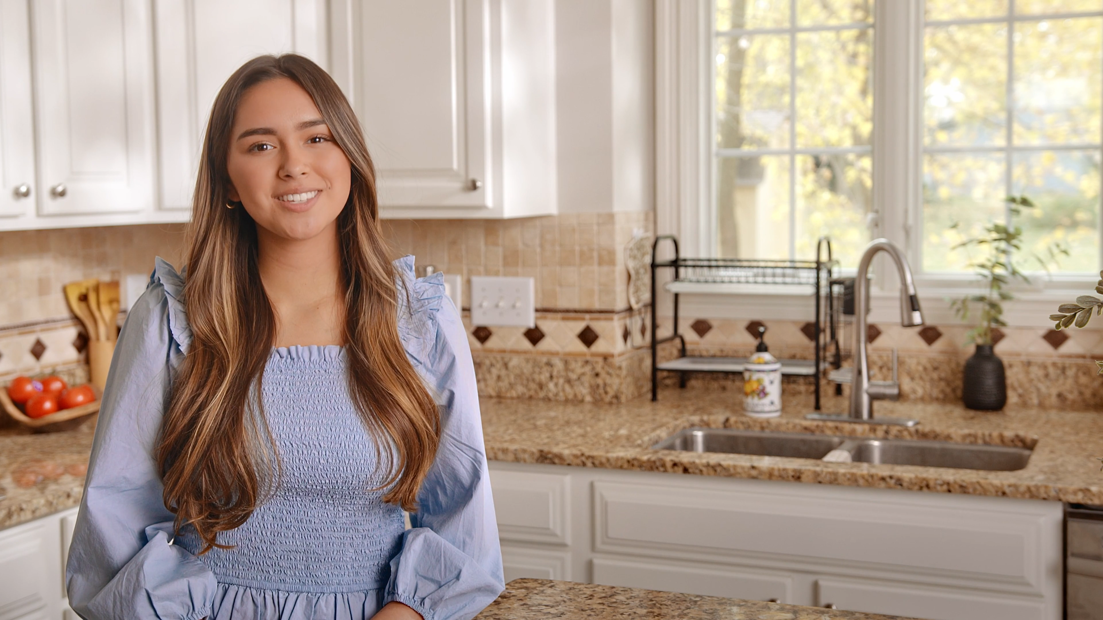 A young woman in a light blue blouse stands in a bright, modern kitchen, smiling confidently at the camera.