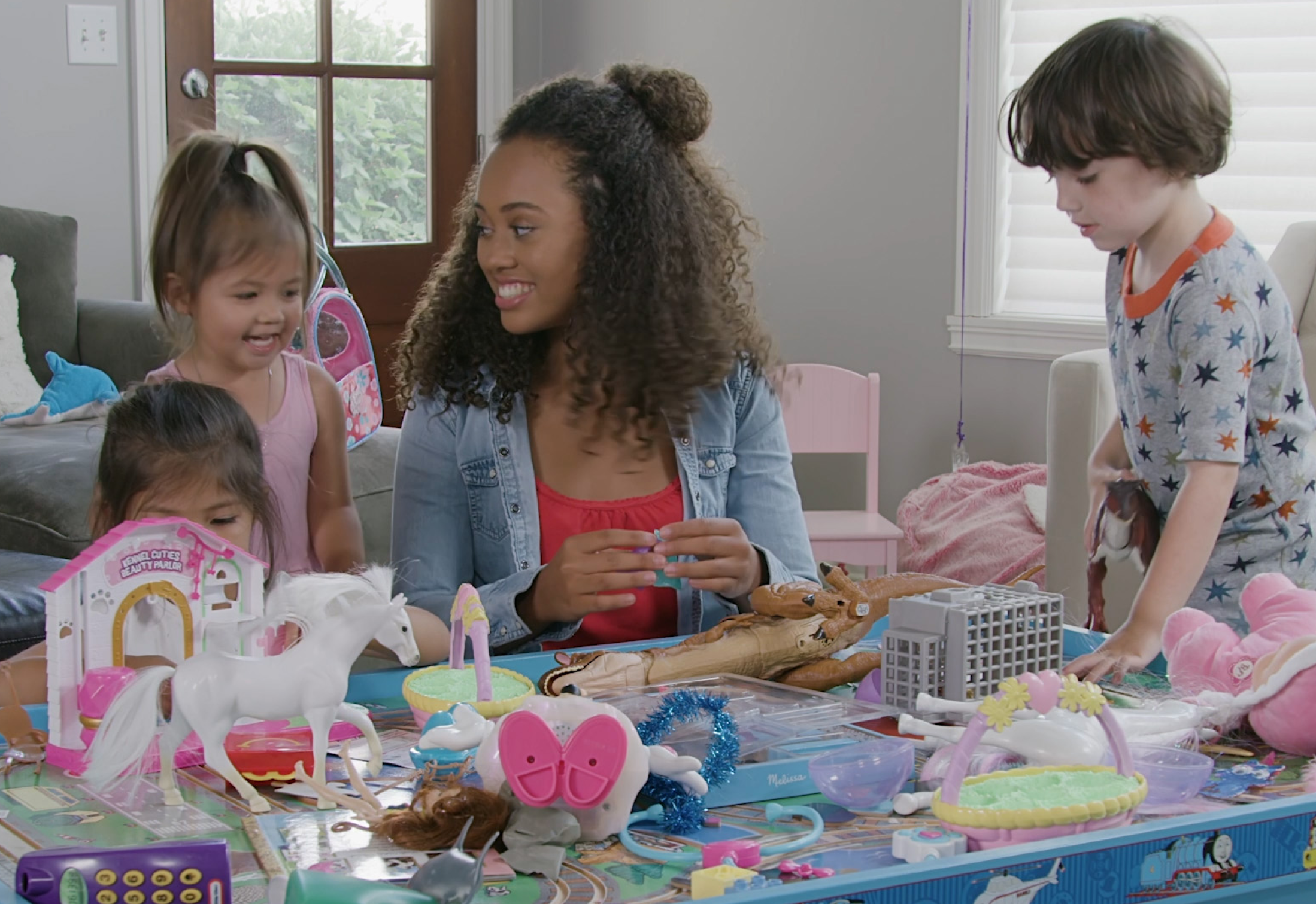 A babysitter and three young children are gathered around a play table filled with toys, including dolls, a toy horse, and a game board. The babysitter, wearing a denim jacket over a red top, smiles while interacting with the children in a well-lit living room.