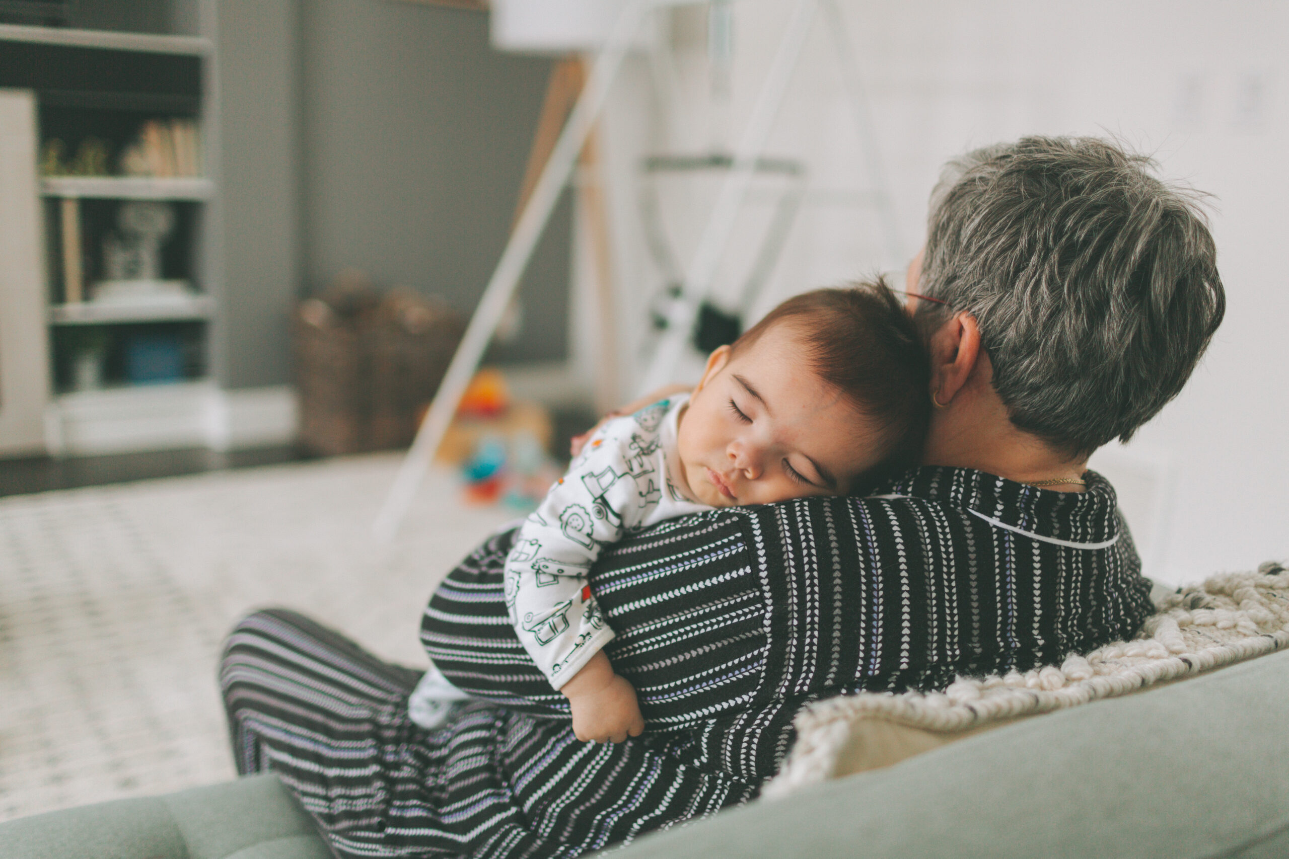 Grandmother holding a sleeping baby on her shoulder while sitting on a couch in a cozy living room.